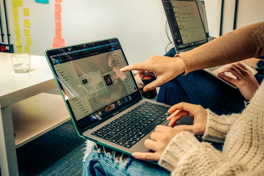 Two people are sitting next to each other, working on laptops. One person is pointing at a laptop screen displaying a digital workspace. Colorful sticky notes are visible on the wall behind them.